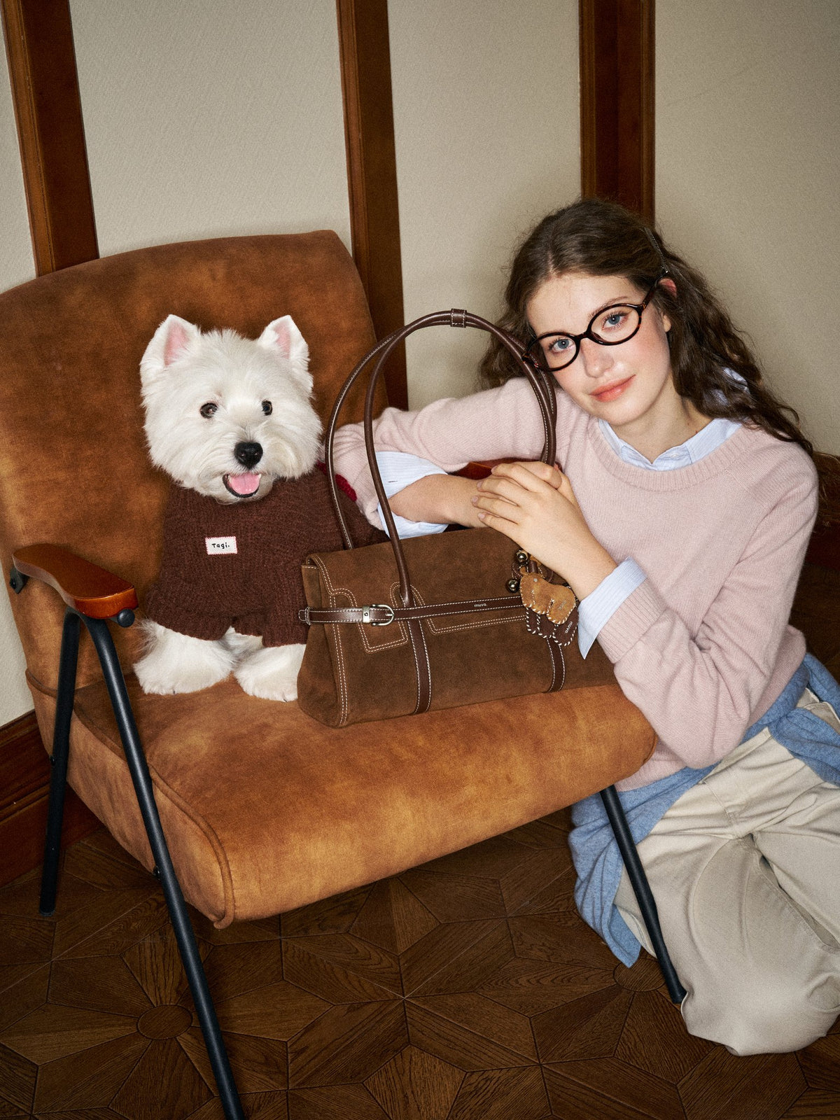 A brown suede muva. shoulder bag on an armchair, beside a white terrier in a knitted jumper